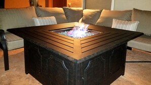 Mesa de fuego cuadrada con un brasero central, rodeada por un armazón de madera oscura, sobre un piso de baldosas.
