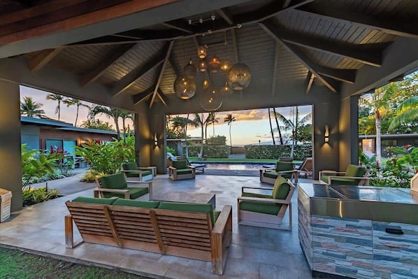 A covered outdoor lounge area with seating, a pool, and a view of palm trees and the ocean.
