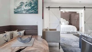A modern bedroom with a bed, a seating area with a chair, a dining table, and a large abstract painting on the wall. - Villa 3 (Kihei)