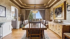 Comedor con mesa de madera, sillas y un candelabro. Se ve una vista a través de la ventana, y la habitación tiene un ambiente rústico con paredes de piedra y piso de madera.