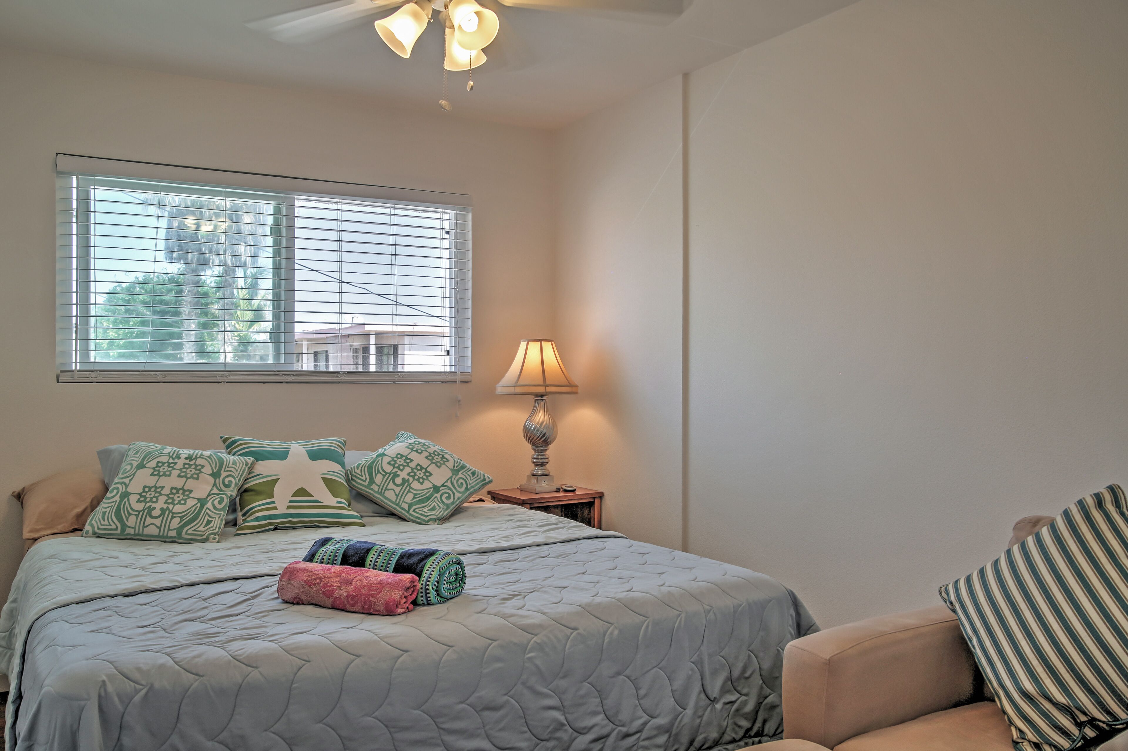 A bedroom with a bed, a nightstand with a lamp, a window with blinds, and a chair.