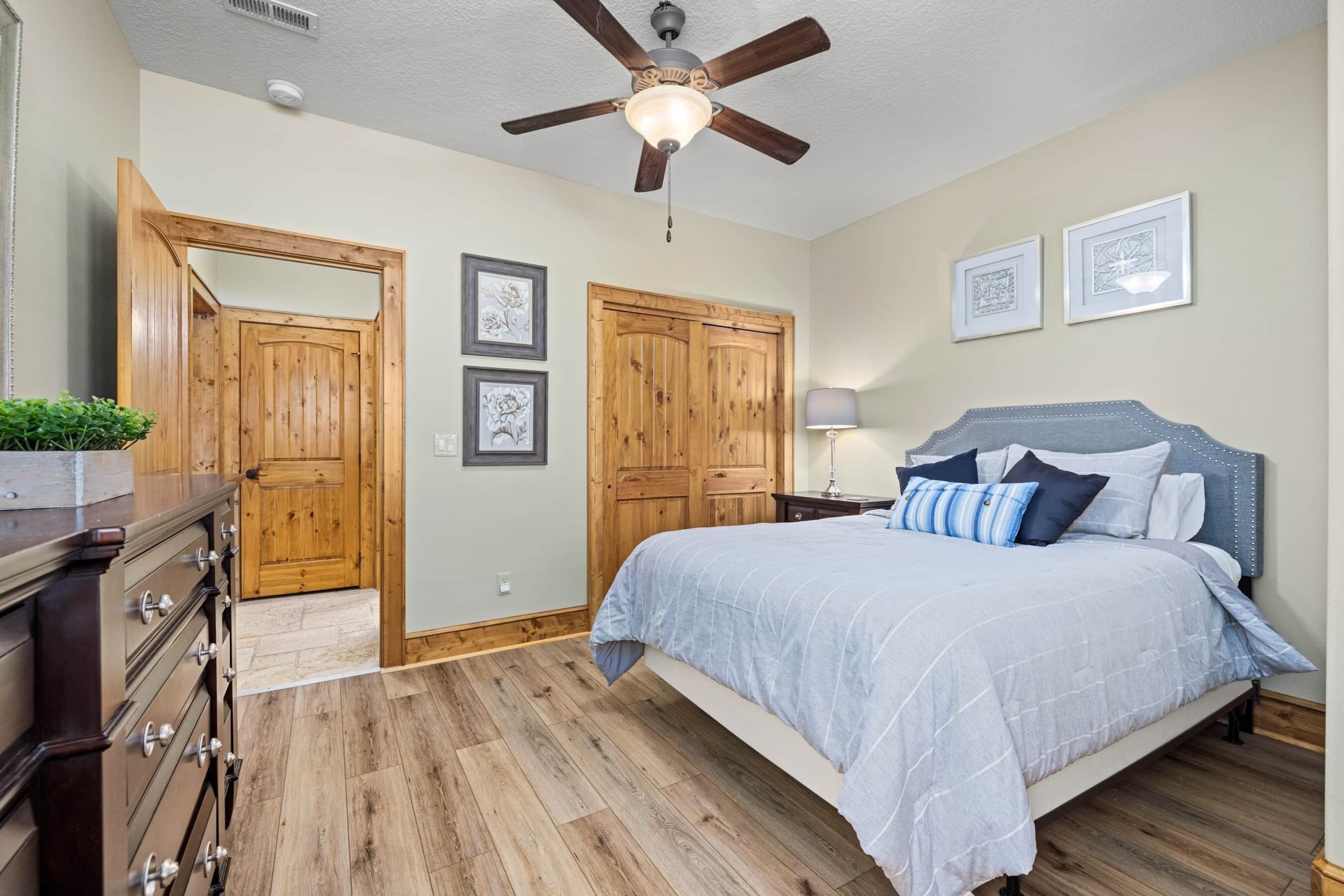 Un dormitorio con suelo de madera, una cama con cubrecama azul, un ventilador de techo y dos puertas de madera.