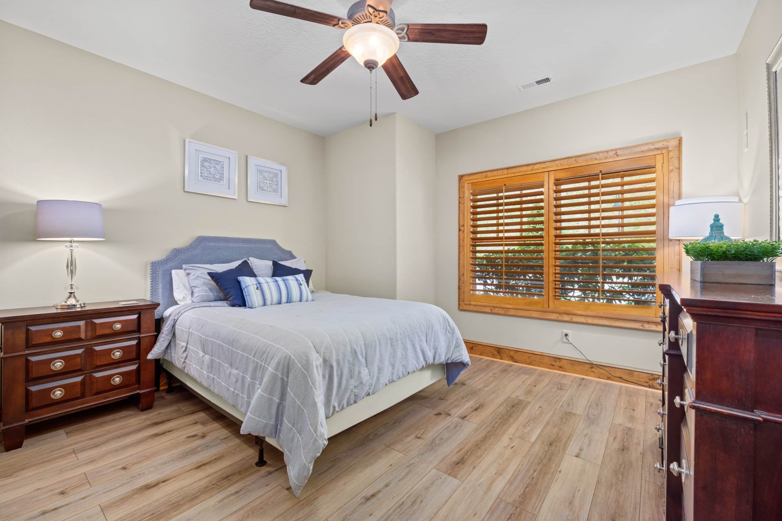 A bedroom with a bed, wooden dresser, nightstand, ceiling fan, window with blinds, and wall-mounted artwork.