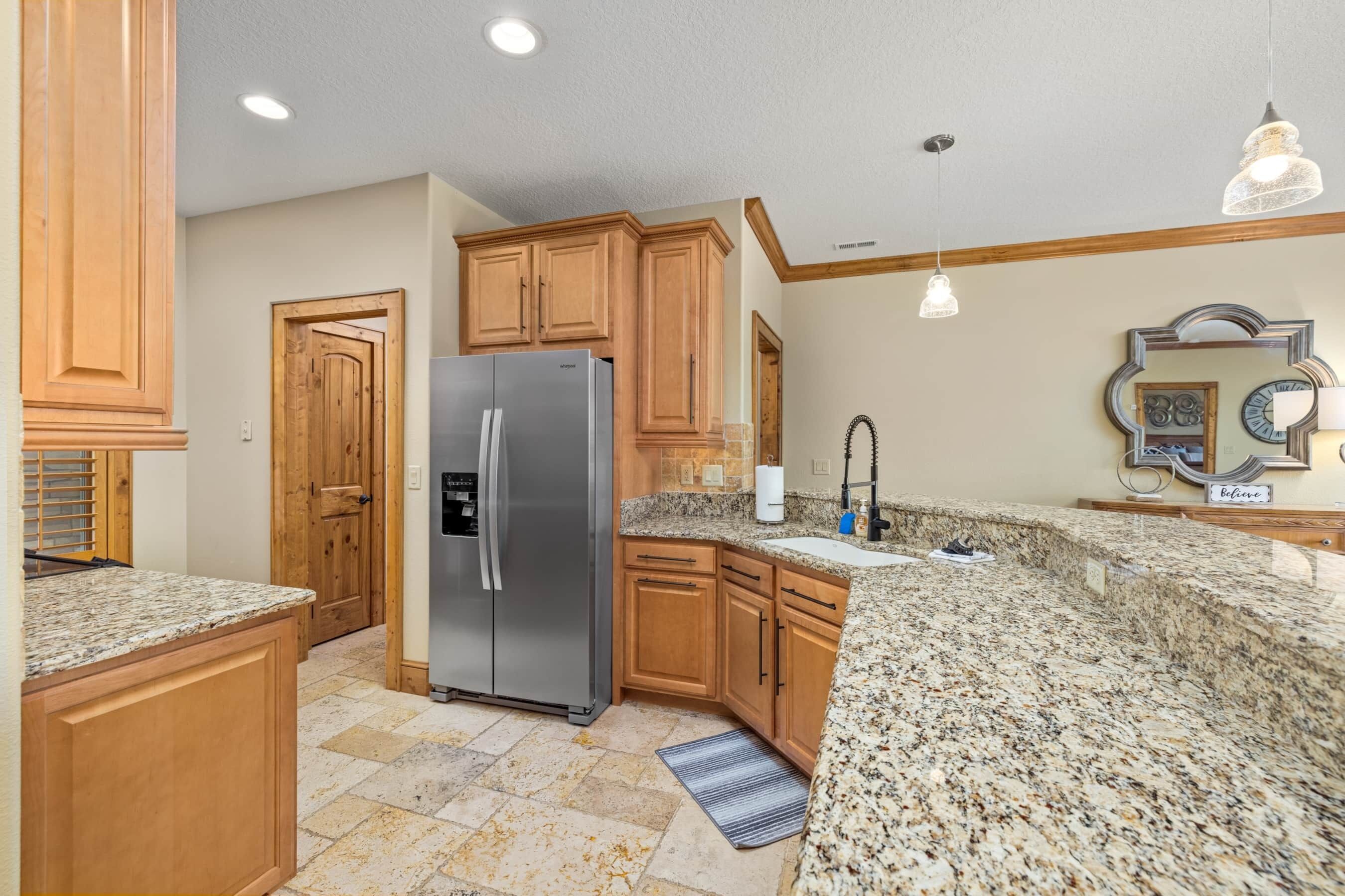 A kitchen with wooden cabinets, granite countertops, a stainless steel refrigerator, and pendant lighting.