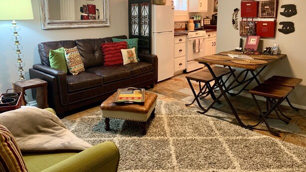 A cozy living room with a brown leather sofa, a green armchair, and a wooden coffee table. A dining area with a wooden table and metal chairs is visible.