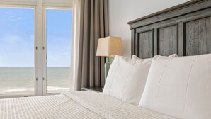 Une chambre avec un grand lit, une table de chevet avec une lampe et une vue sur l’océan par une porte coulissante.