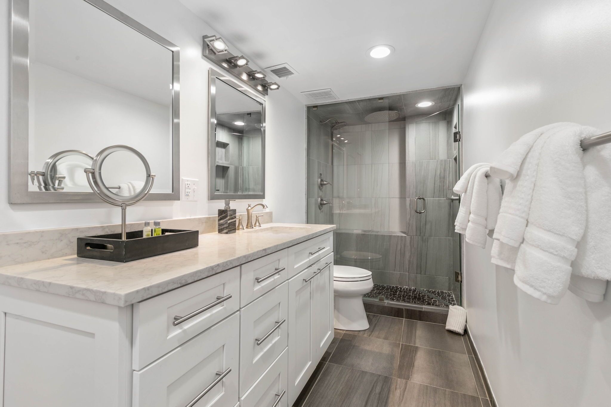 A modern bathroom with a marble countertop, a large mirror, a glass-enclosed shower, and white towels.