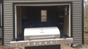 A gas grill in front of a garage with a dark gray exterior and a triangular roof.