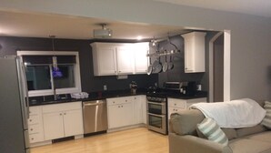 A compact kitchen with white cabinets, a stainless steel dishwasher, a stove, and a microwave. There's a beige sofa with a blanket and pillows, and a window with blinds.