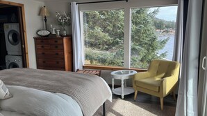 Une chambre à coucher comprenant un lit, une chaise, une petite table, une commode, une lampe et une fenêtre donnant sur des arbres et une étendue d’eau.