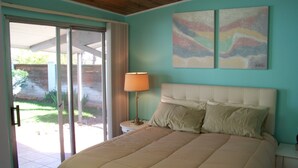 A bedroom with a bed, a nightstand, a lamp, a sliding glass door, and two abstract paintings on the wall.