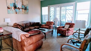 Una sala de estar con un sofá de cuero marrón, un sillón estampado, una silla de madera y una mesa de centro de vidrio. Hay un cuadro en la pared, una lámpara en una mesita auxiliar y una ventana con vista a un puente.