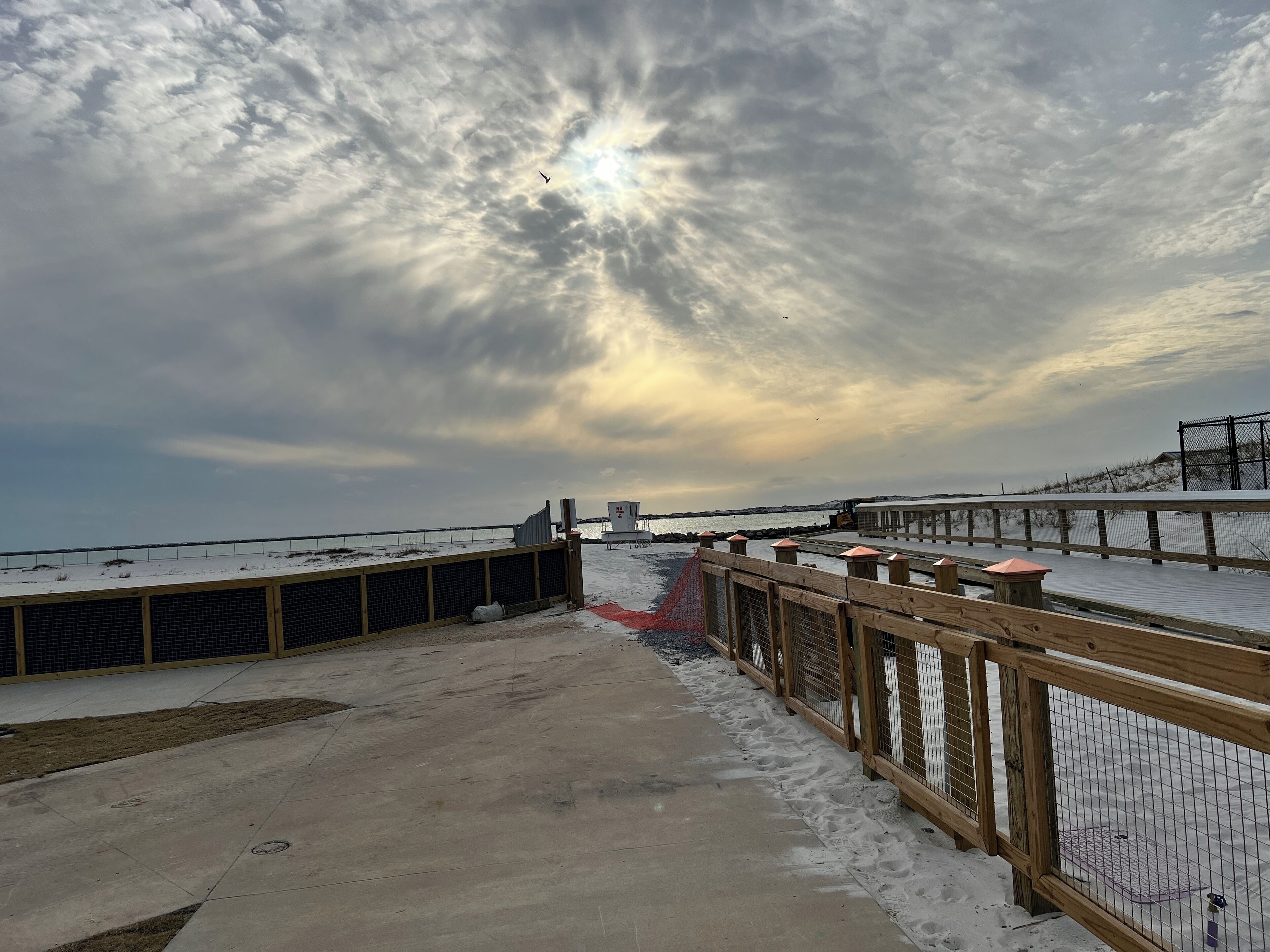 A snowy deck with a wooden railing, a concrete pathway, and a view of the sea under a cloudy sky.