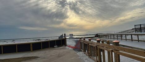 A snowy deck with a wooden railing, a concrete pathway, and a view of the sea under a cloudy sky.