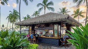 Een bar met rieten dak en een stenen vloer, omringd door tropische planten en palmbomen.