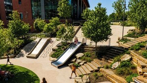 Un'area giochi con scivoli e scalinate, circondata da alberi e un edificio sullo sfondo.