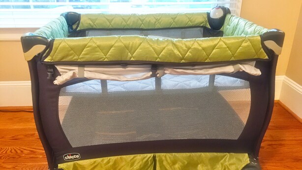 A portable playpen with a green and black color scheme, placed on a wooden floor near a window.