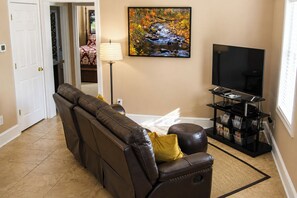 A living room with a brown leather sofa, a flat-screen TV, a lamp, and a painting of a stream.