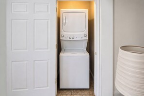 A laundry room with a front-loading washer and dryer, a white door, and a cylindrical container. - The Cary Cottage 1 BR 1 BA Private Sleeps 4 - Downtown Cary, Eat Drink Play Shop (Cary)