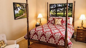 Una cama con dosel con un edredón de estilo damasco rojo y blanco, dos lámparas de noche, una mesita de noche con lámpara y una silla con una manta.