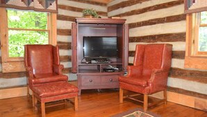 Interior de una cabaña de troncos con dos sillones de cuero, un mueble de entretenimiento de madera y un ventanal grande.