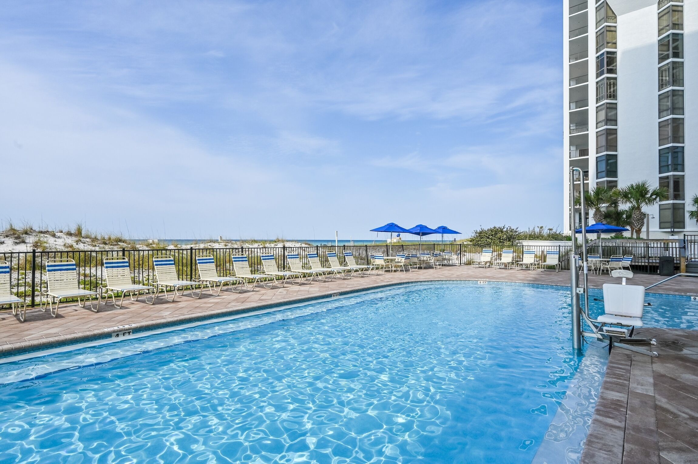 A swimming pool with lounge chairs and umbrellas, surrounded by a fence and a building.