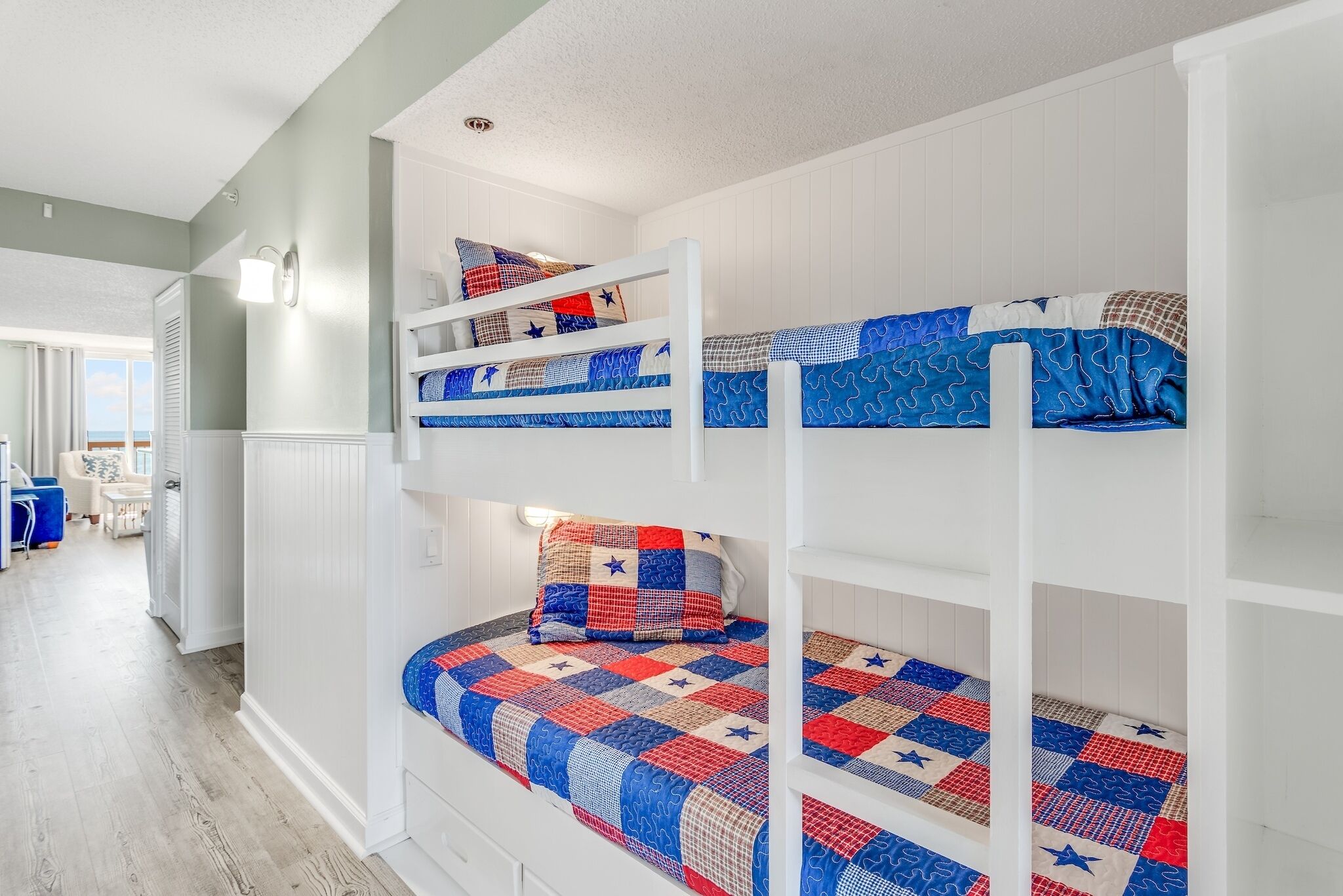 A bunk bed room with a checkered bedspread and pillows, white walls, and a hallway leading to other rooms.
