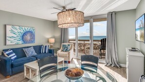 A modern living room with a glass dining table, a blue sofa, and a view of the ocean. - Spectacular Views,  Oceanfront Condo, Renovated (Destin)