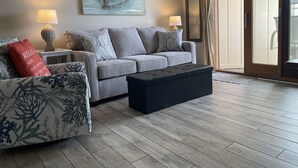 A living room with a grey sofa, a black ottoman, and a painting of boats on the wall. - Beachfront/Oceanfront Hilton Head Island Condo (Hilton Head)