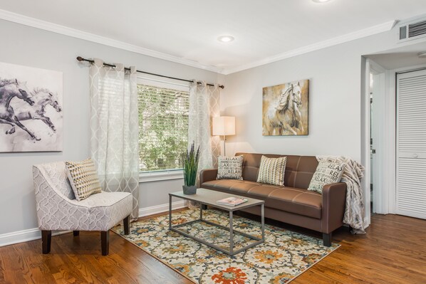 Una sala de estar con un sofá de cuero marrón, un sillón estampado, una mesa de centro y un ventanal con cortinas traslúcidas.