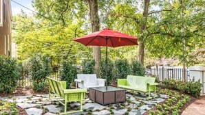 Un patio al aire libre con un paraguas rojo, sillas verdes y blancas, y una mesa cuadrada.