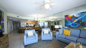 A spacious living room with a blue sofa, two armchairs, and a coffee table. A kitchen is visible in the background with wooden cabinets and a dining area. - Phoenix10 Beach front condo fully upgraded with brand new custom bathrooms. (Orange Beach)