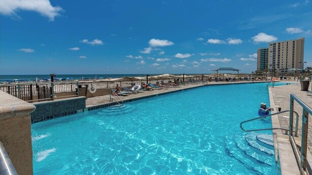 A swimming pool with clear blue water, surrounded by lounge chairs and umbrellas, with a beach and ocean in the background. - Phoenix10 Beach front condo fully upgraded with brand new custom bathrooms. (Orange Beach)