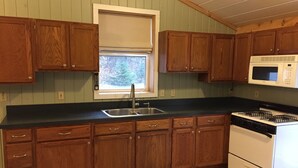 A kitchen with wooden cabinets, a stainless steel sink, a white stove, and a window with a white blind. - Cozy and comfortable 1,000 sq. ft. 2-bedroom home with garage. (Bellaire)