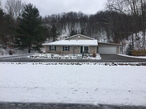 A single-story house with a snow-covered roof and driveway, surrounded by trees and a clear sky.