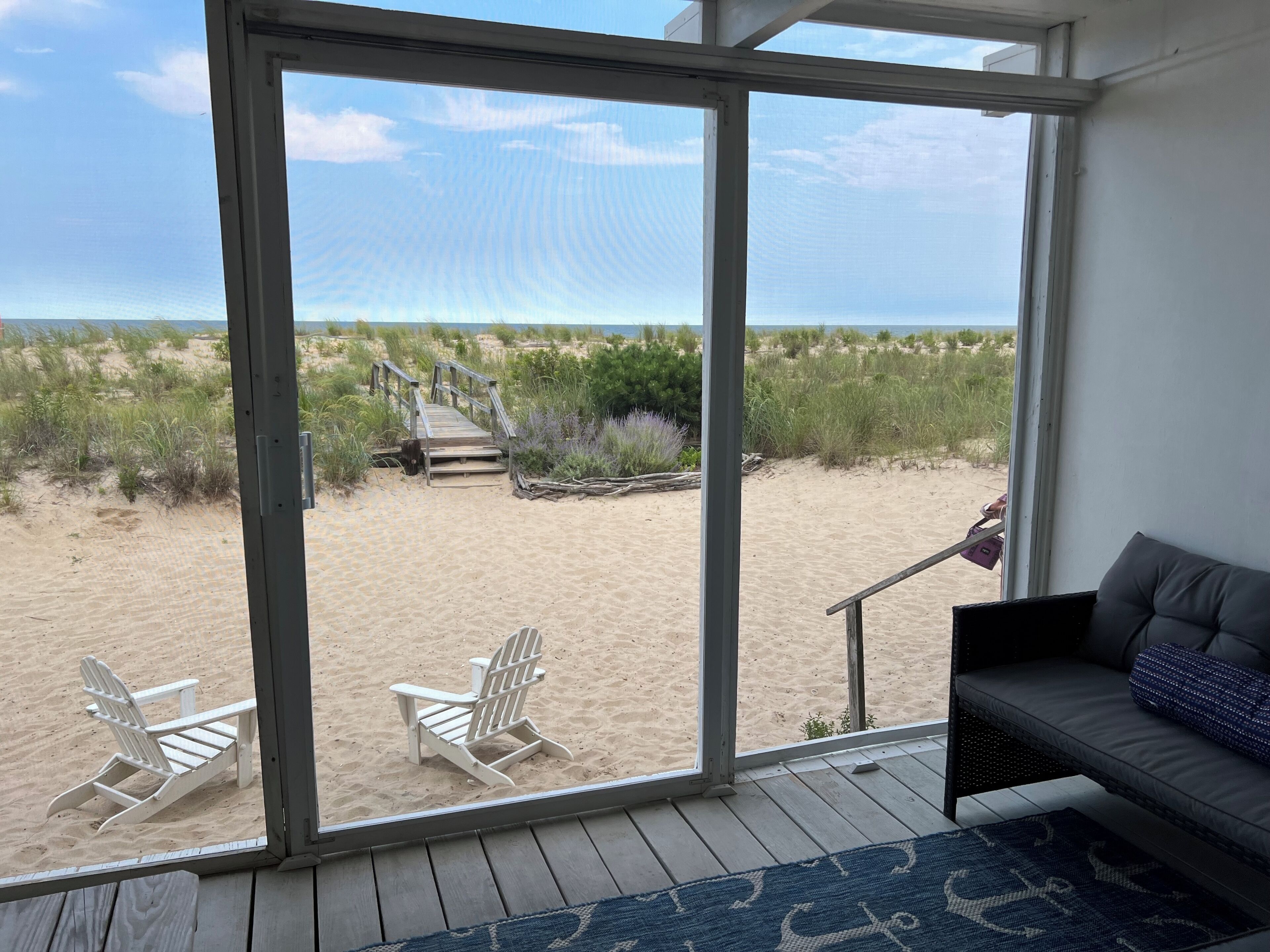 Uitzicht op het strand vanuit een kamer met een grote schuifglazen deur, een grijze bank en twee witte strandstoelen.