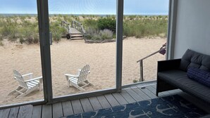 Strandudsigt fra et værelse med en stor skydedør, en grå sofa og to hvide strandstole.