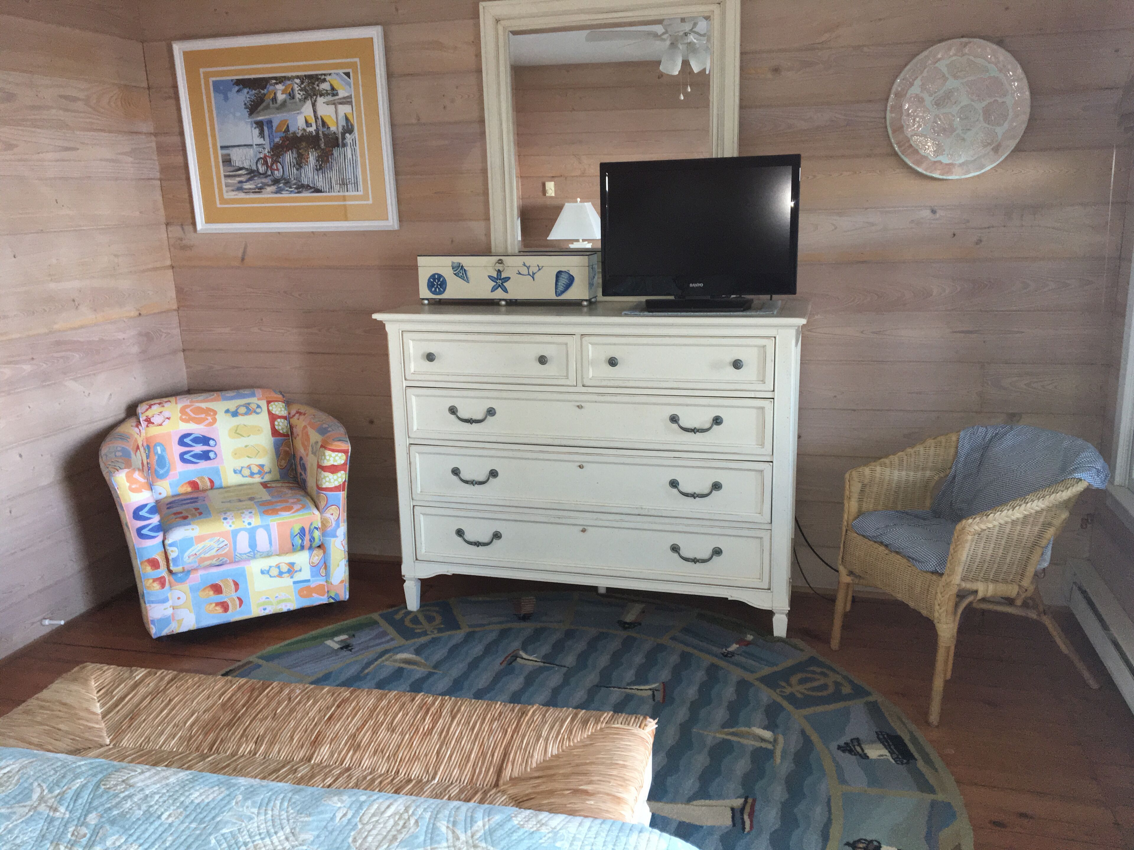 Una habitación con un aparador blanco, un televisor encima, una silla colorida, una silla de mimbre y una alfombra estampada.