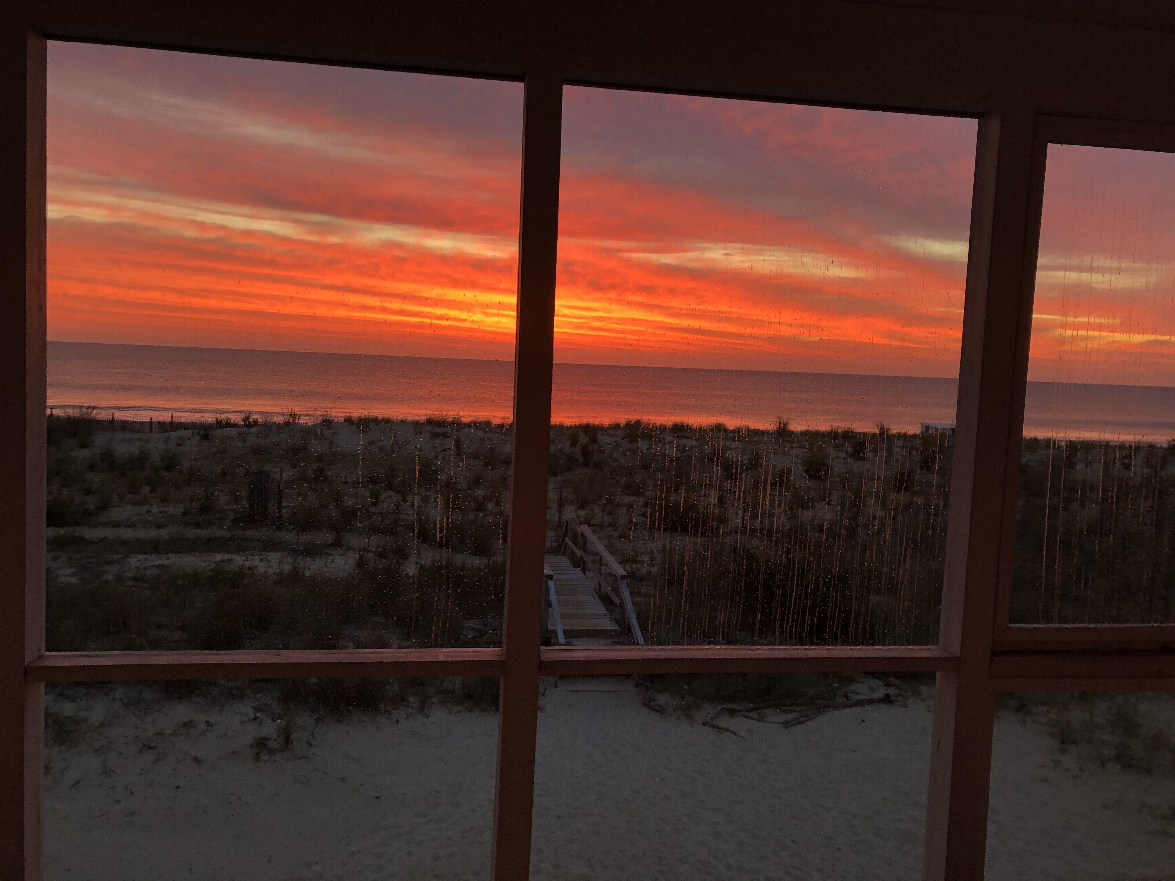 Zonsondergang te zien door een raam met houten kader, zandstrand en een pad dat naar het water leidt.
