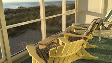 Een kamer met grote ramen met uitzicht op een strand. Twee Adirondack-stoelen met gestreepte kussens en een houten tafel.
