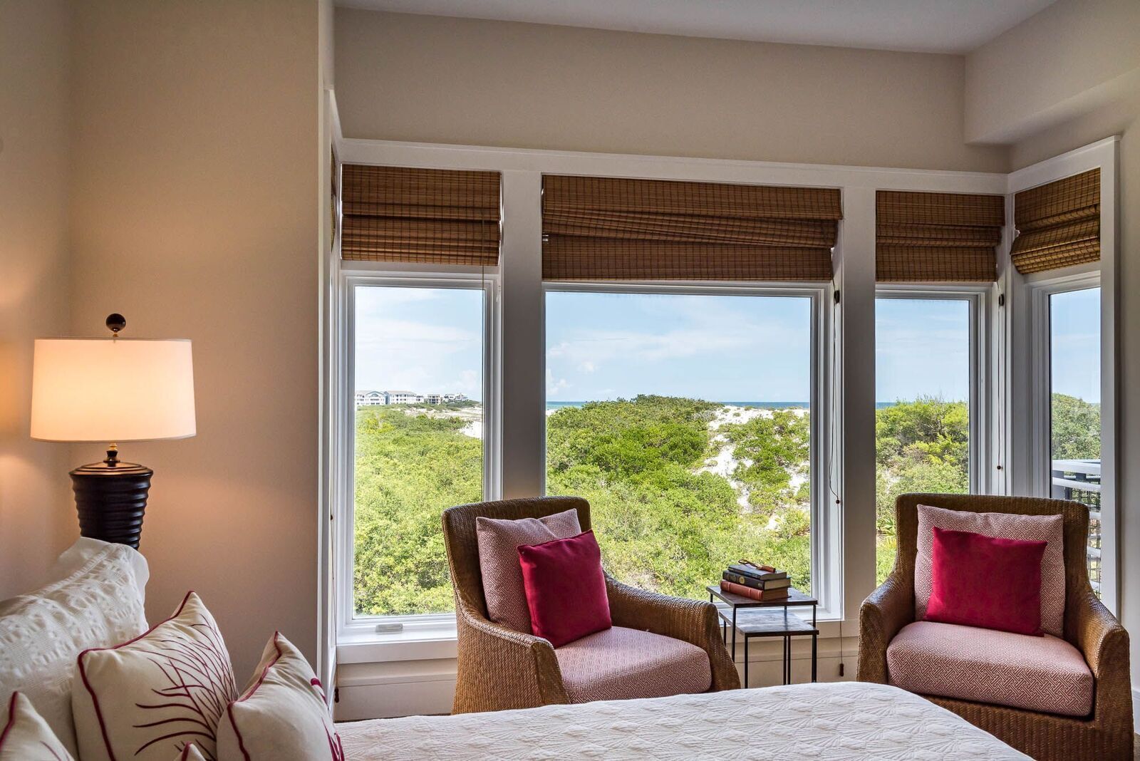 A hotel room with a bed, two wicker chairs with red cushions, a small table with books, and large windows with blinds.