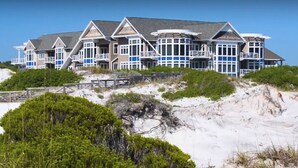 A large, multi-story beachfront property with a prominent balcony area and a wooden deck.