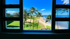 A view through a window with a beach and palm trees. - Serene Ocean-Front Condo at Menehune Shores - Unit 520 - Steps from the Ocean! (Kihei)