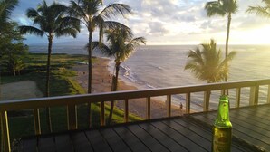 A balcony with a view of the beach and ocean, a bottle of beer, and palm trees. - Serene Ocean-Front Condo at Menehune Shores - Unit 520 - Steps from the Ocean! (Kihei)