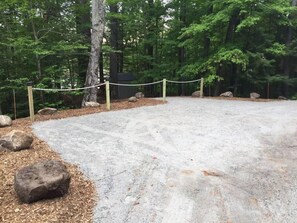 Une aire de stationnement en gravier, délimitée par une barrière en corde et des rochers, et entourée d’une zone boisée.