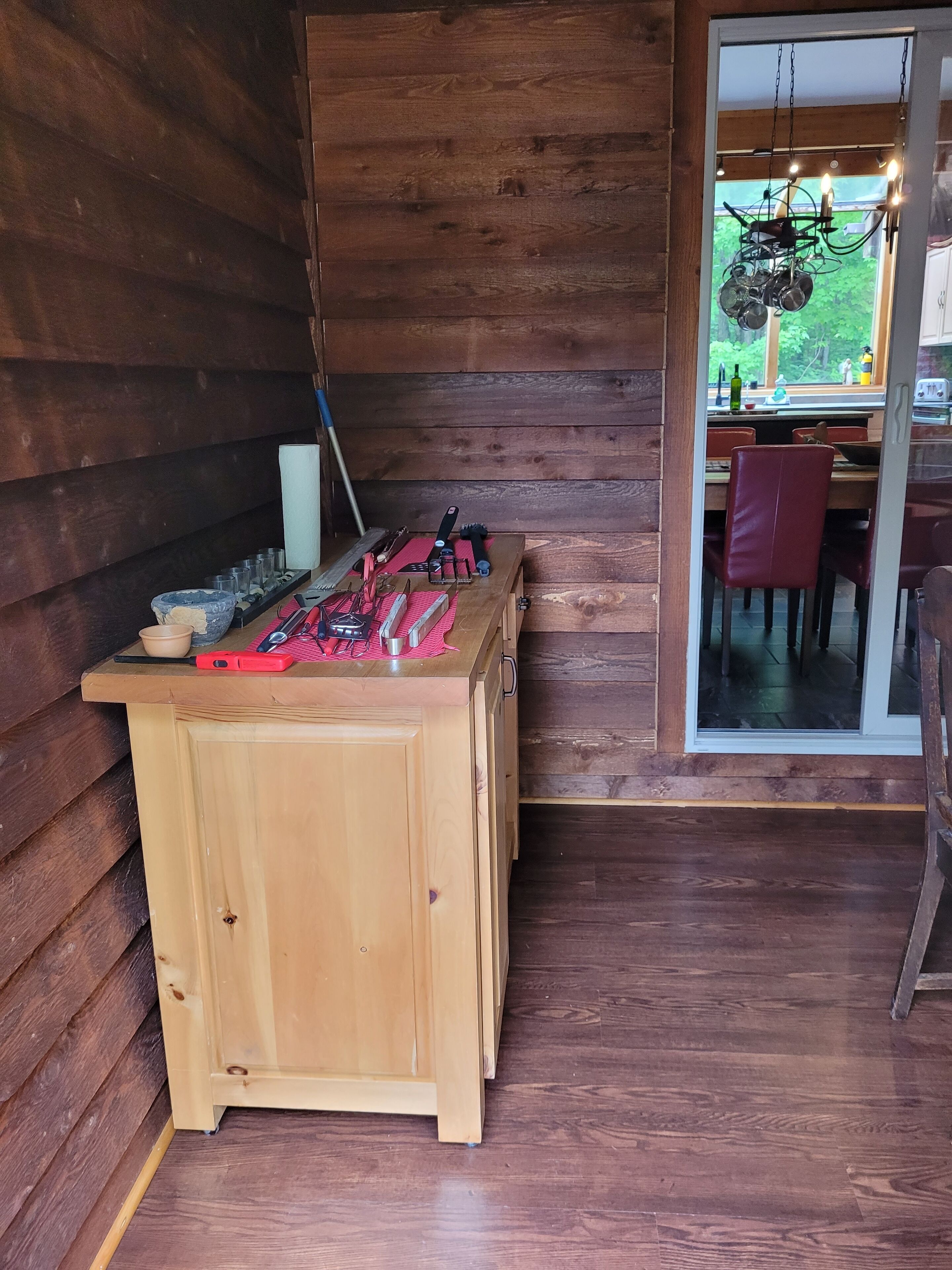 Une table en bois avec des outils et un tissu rouge, une fenêtre donnant sur un paysage verdoyant, et un coin repas avec des chaises et une table.