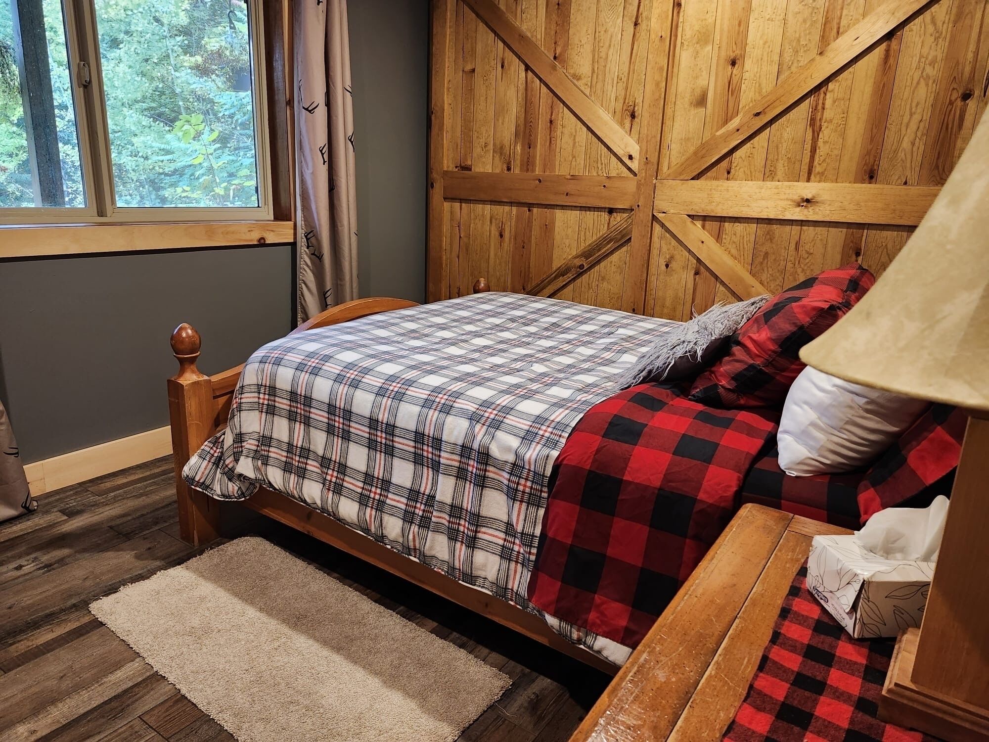 A bedroom with a wooden bed, plaid bedding, a nightstand, a lamp, and a window with curtains.