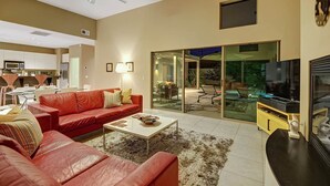 A modern living room with a red leather sofa, a coffee table, and a flat-screen TV. There is a sliding glass door leading to a patio area with outdoor furniture. - Cool Modern Pool & Spa, 2 Bedroom + Private Den House. (Palm Springs)