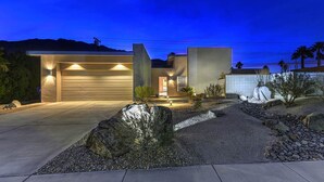 A modern house with a garage, well-lit entrance, and landscaped garden with rocks and plants. - Cool Modern Pool & Spa, 2 Bedroom + Private Den House. (Palm Springs)