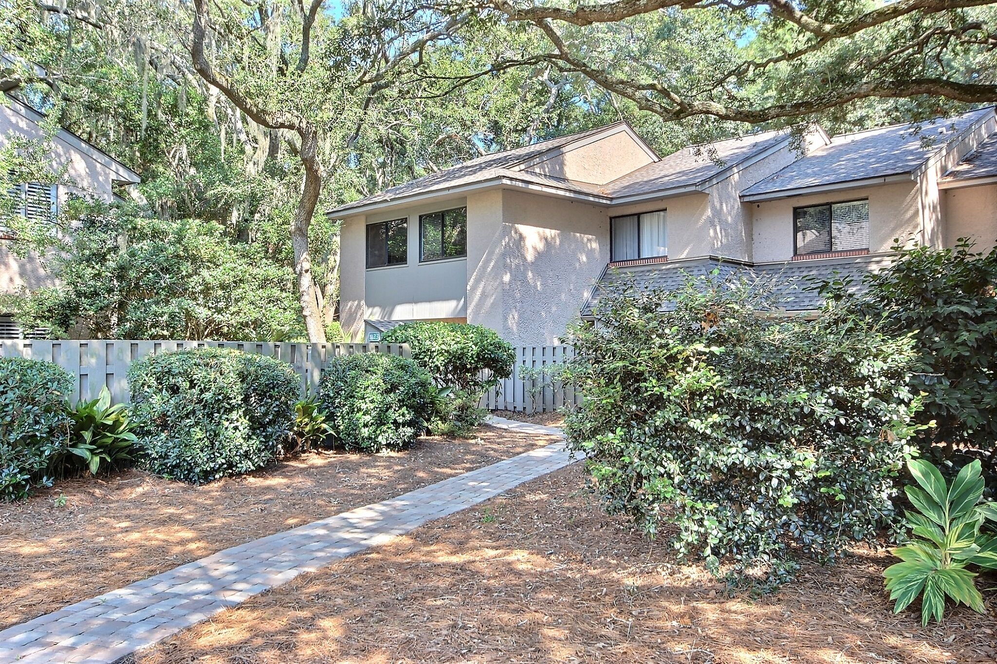 A residential property with a paved driveway, a wooden fence, and various shrubs and trees.